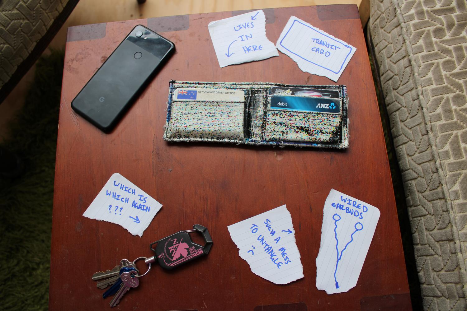 An aerial photo of a table containing items I carry each day, with pieces of paper beside each item describing the problems with each. The photo contains my phone, wallet, keys and wired earbuds. I didn't actually have the wired earbuds anymore so I've just drawn a picture to represent them.