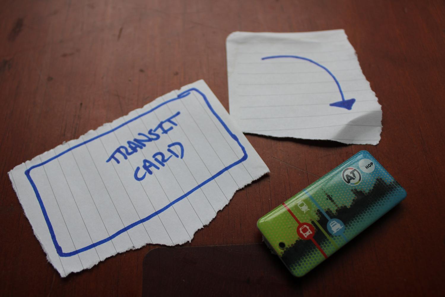 A close up photo of a table surface containing three items: A piece of paper representing my physical transit card, an arrow pointing to the right and a smaller, badge sized transit card designed for keychains. The arrow is supposed to symbolise that I've gone from one transit card to another.