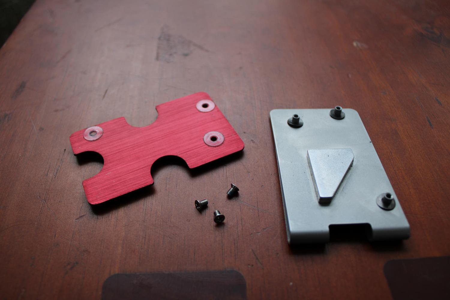 A close up photo of a table surface containing a metal device that has been unscrewed. The base plate is on the right with three little stands to provide room in between itself and the top plate. The idea is that the cards sit in between the gap. The top plate is red and sitting to the left of the base plate with three small screws sitting just below it.