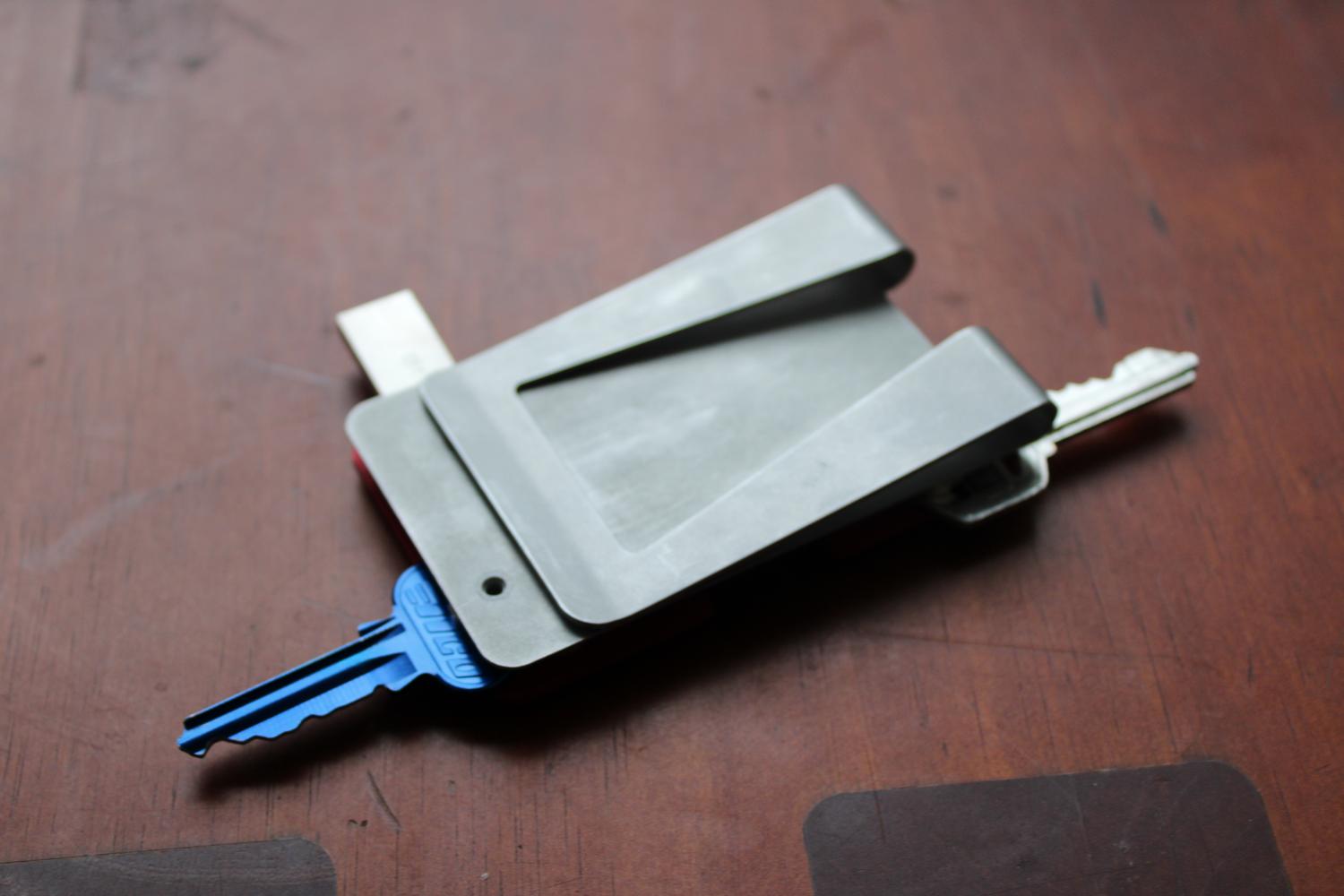 A close up photo of a table surface with the assembled metal wallet sitting on it. The wallet is upside down, with the keys and USB drive extended beyond the base plate. The bottom of the base plate is visible, as the wallet is upside down, and has a large clip at the bottom for slotting cards into.