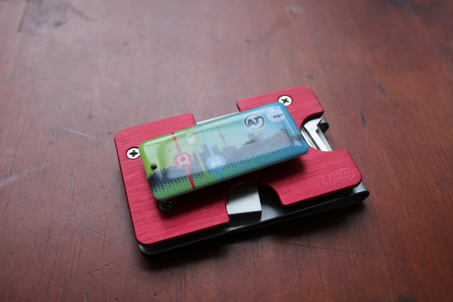 An aerial photo of the assembled metal wallet sitting on a table surface. It is positioned right side up this time showing the red top plate. On top of the red plate is the small badge sized transit card, seemingly hovering in place. It's actually attached to a velcro strip although you can't visually tell that from the photo.