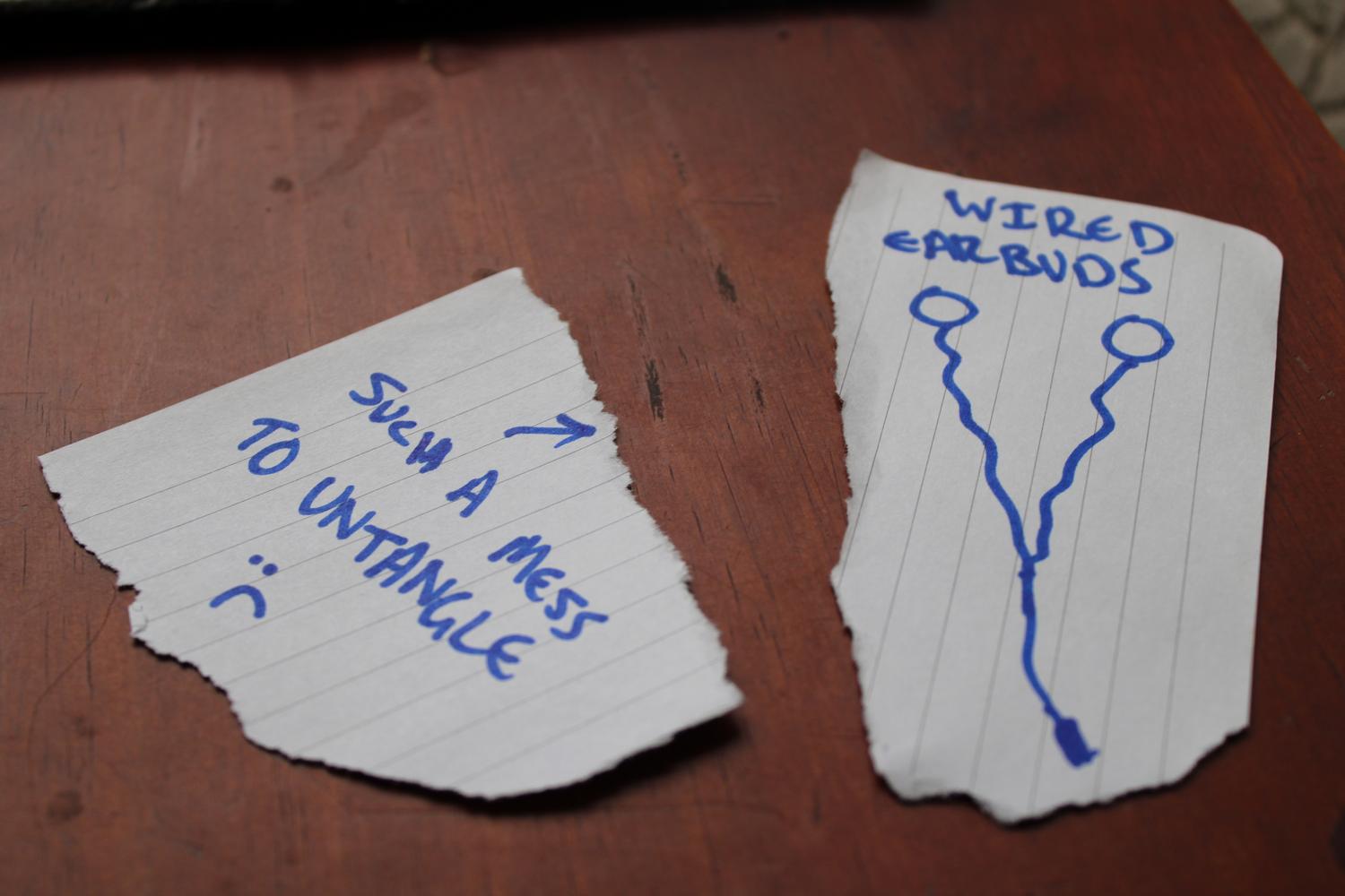 A close up photo of a table surface with two pieces of paper. One on the left says "Such a mess to untangle" with a sad face and an arrow pointing to the right. The piece of paper on the right is a ridiculously bad drawing of wired earbuds with text at the top that reads "Wired earbuds". I no longer had the them in my position so this is supposed to be a placeholder for the real thing.
