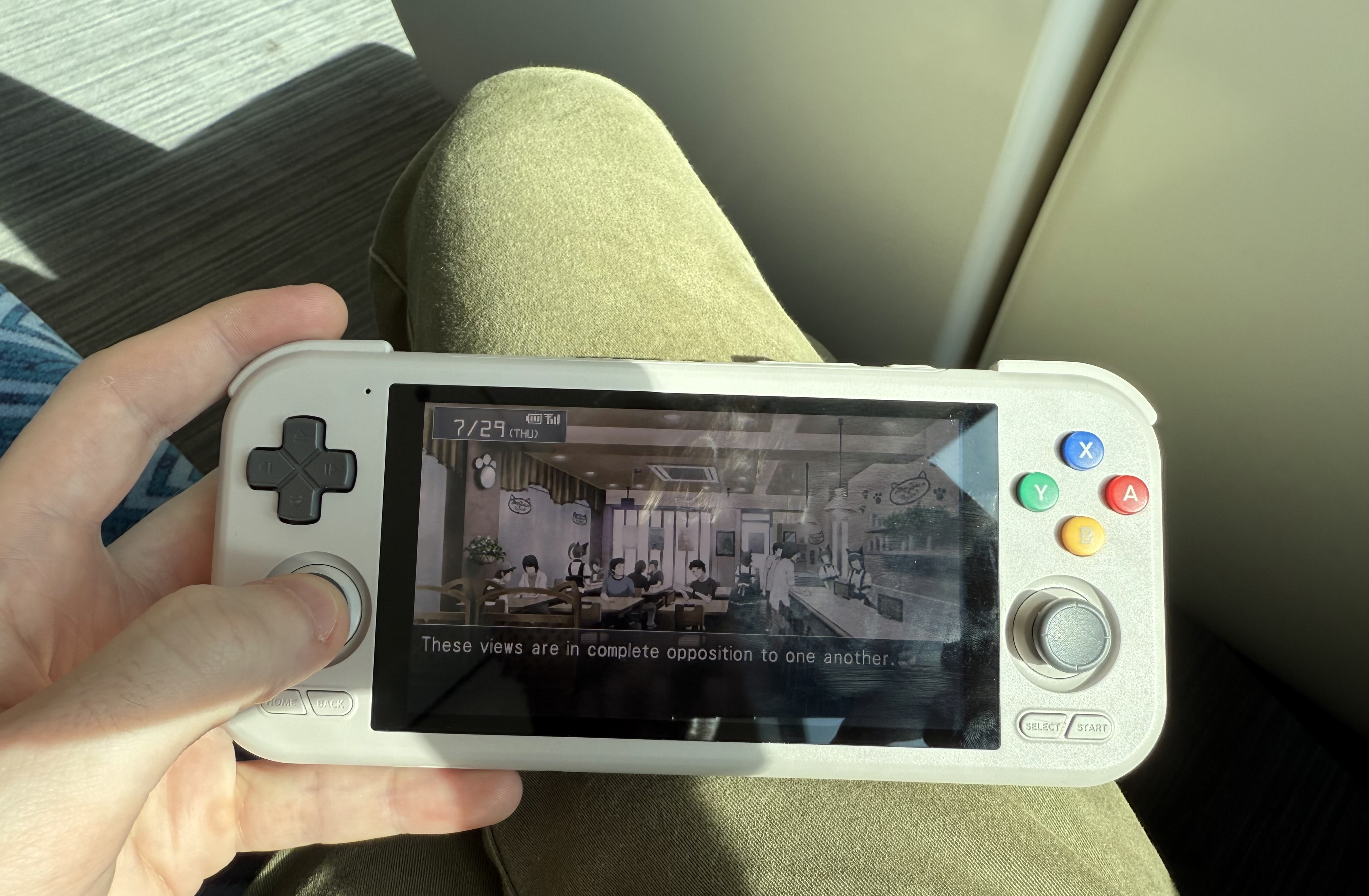 A photo of the author holding a Retroid Pocket 4 Pro with their left hand. Visible on the screen is Steins;Gate for the Playstation Portable. It's not entirely clear from the image but they are on a train.